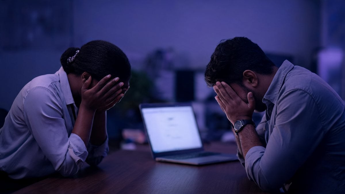 Two exhausted colleagues at a laptop in a dim purple-lit room