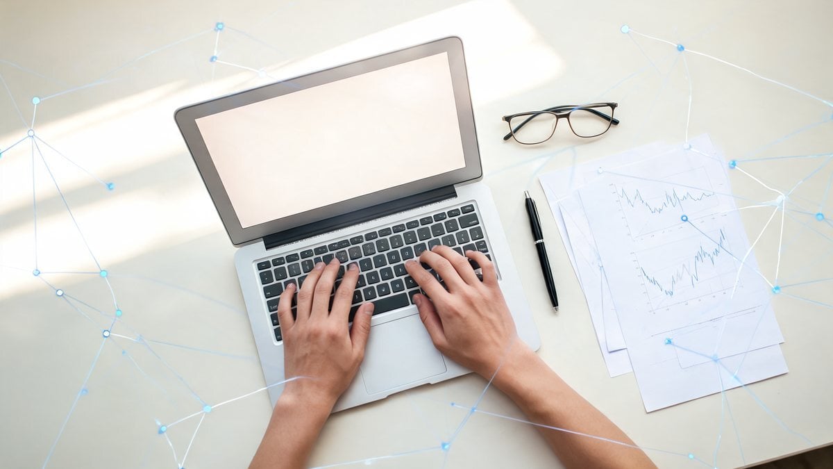 Hands typing on a laptop beside glasses, a pen and chart printouts