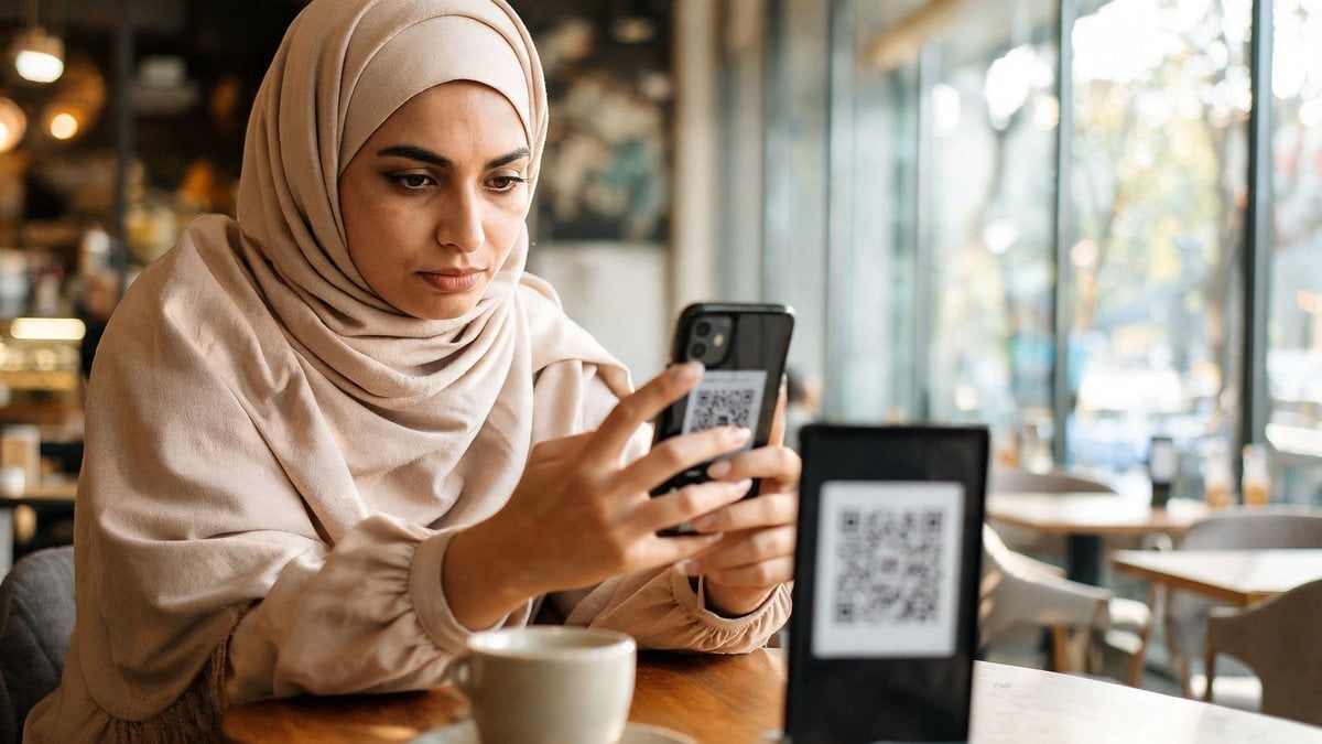 A woman wearing a hijab in a cafe, scanning a QR code on a table stand with her smartphone